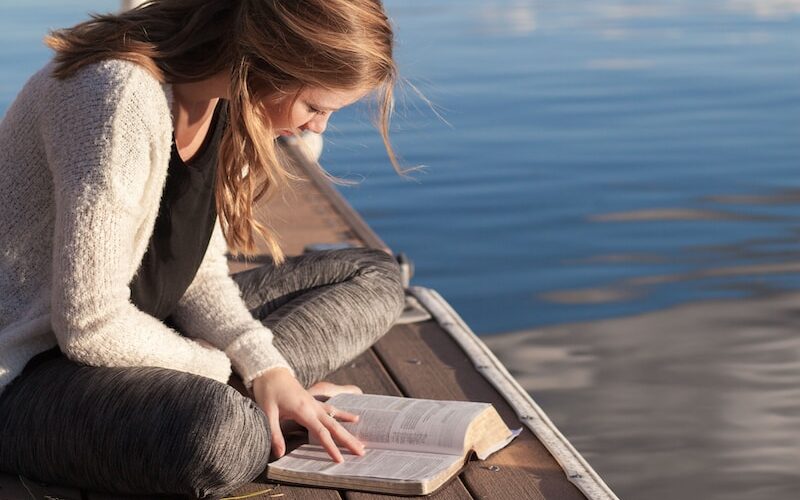 photo of woman reading book near body of water