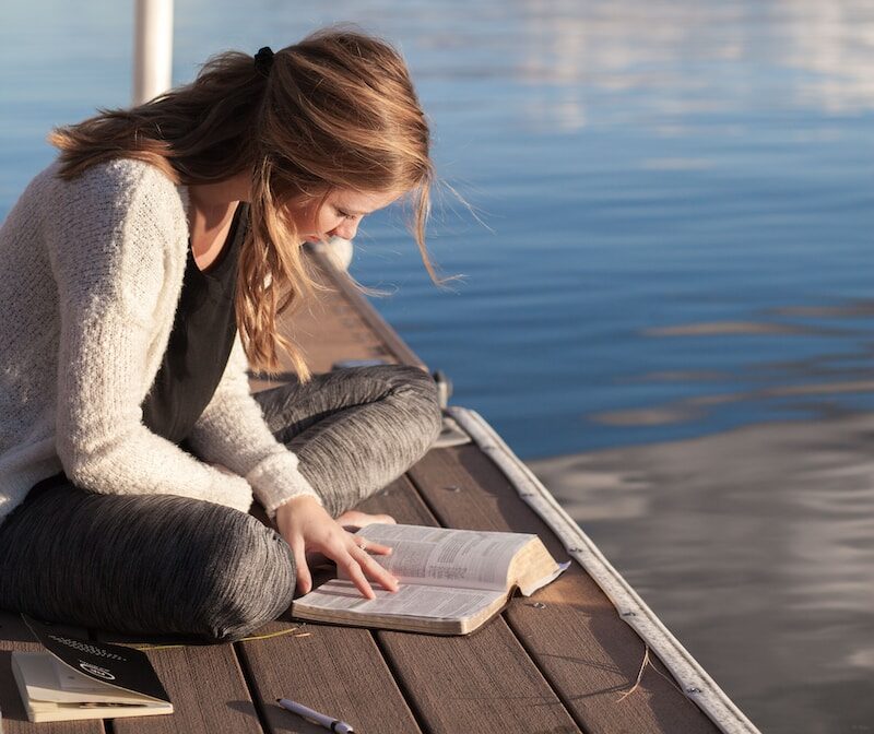 photo of woman reading book near body of water