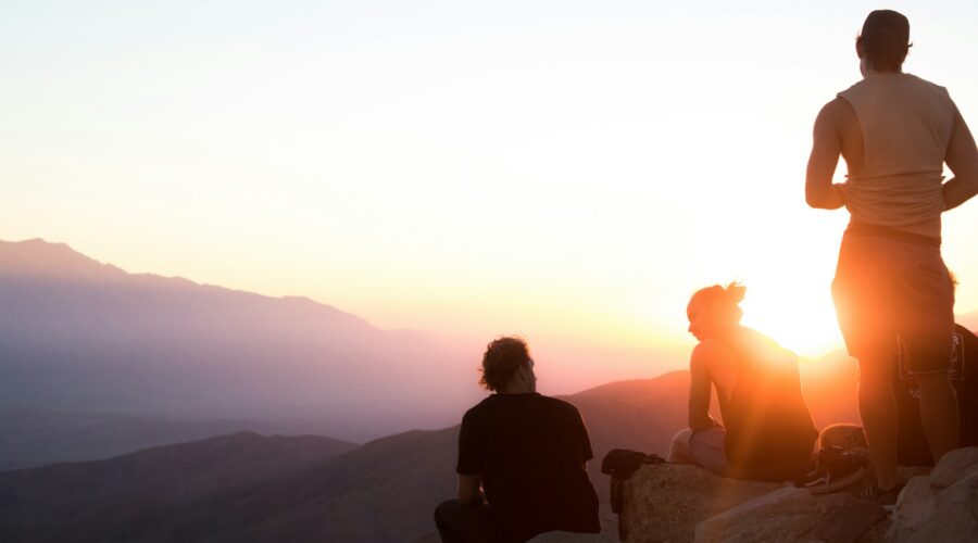 two men sitting and one man standing near cliff taken during golden hour